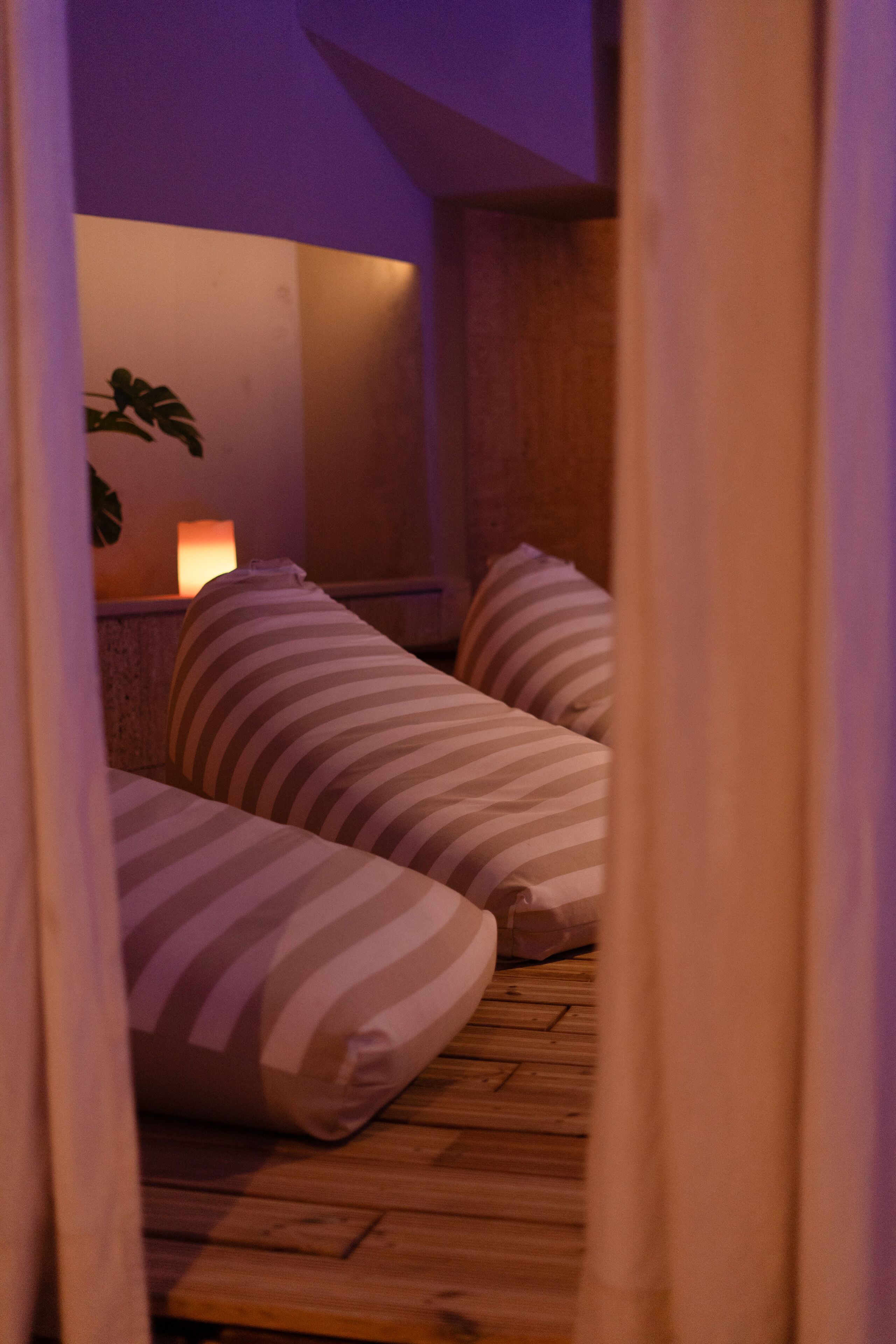 A dimly lit room with a wooden floor, striped pillows, and a wall-mounted lamp.
