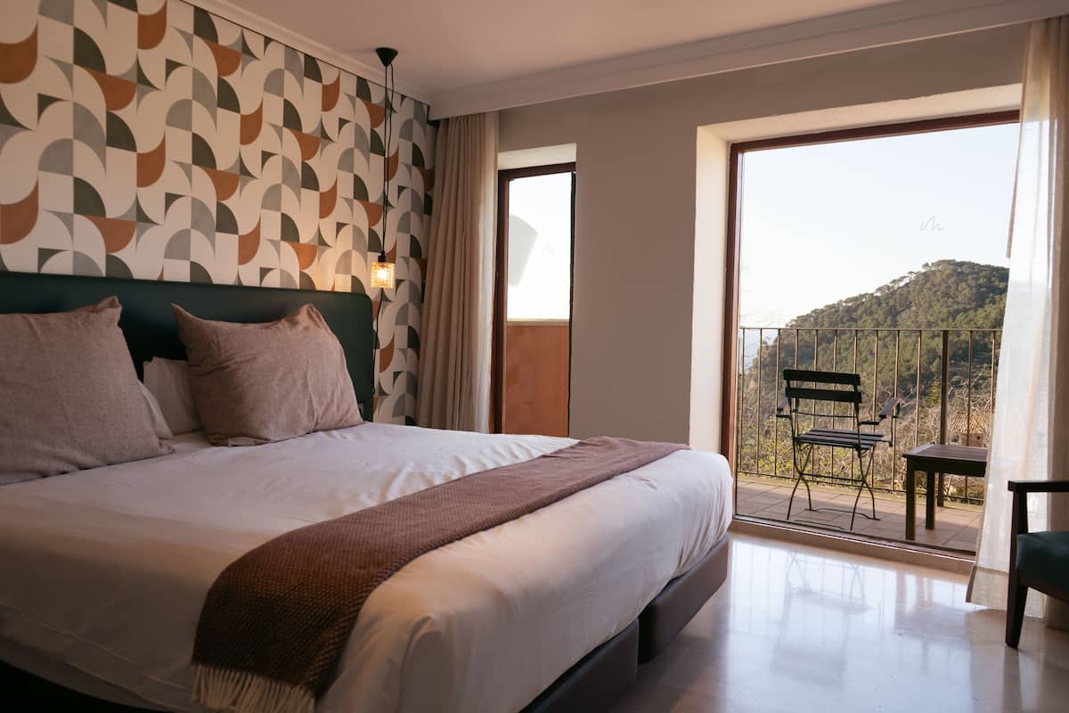 A hotel room with a large bed, a balcony with a chair, and a view of a hill.