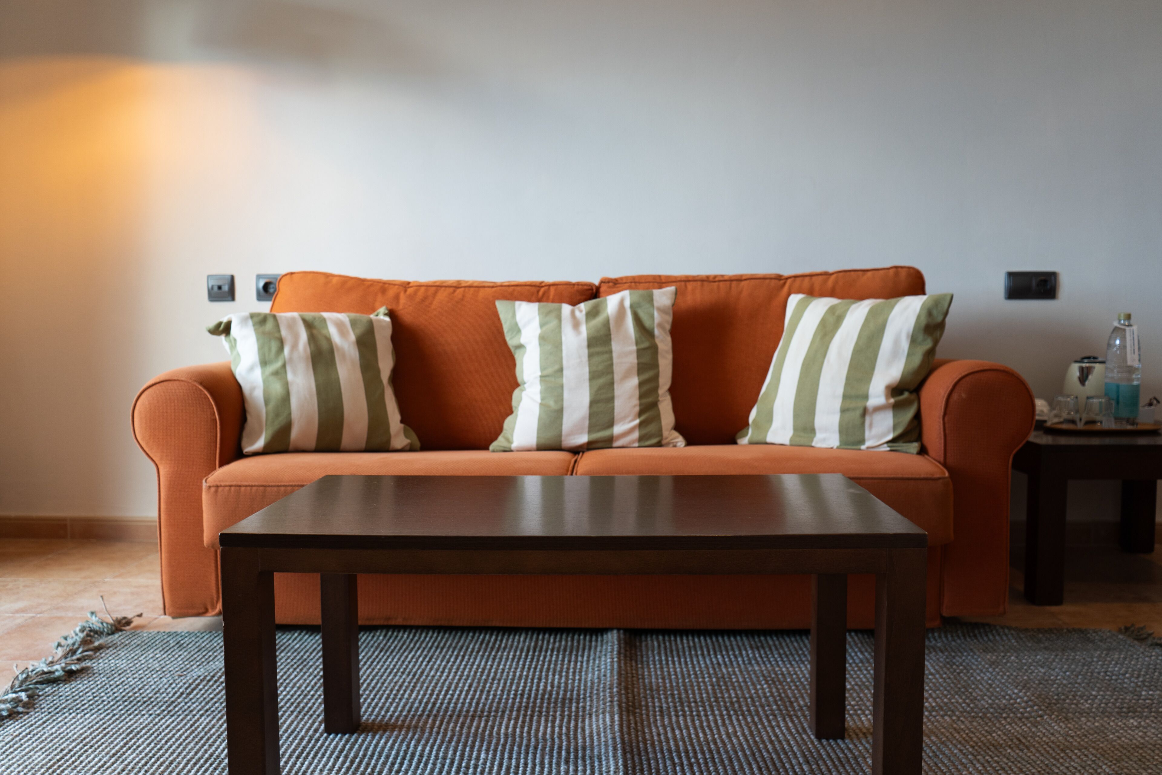 Una habitación con un sofá naranja, dos cojines a rayas, una mesa de centro de madera oscura y una alfombra estampada.