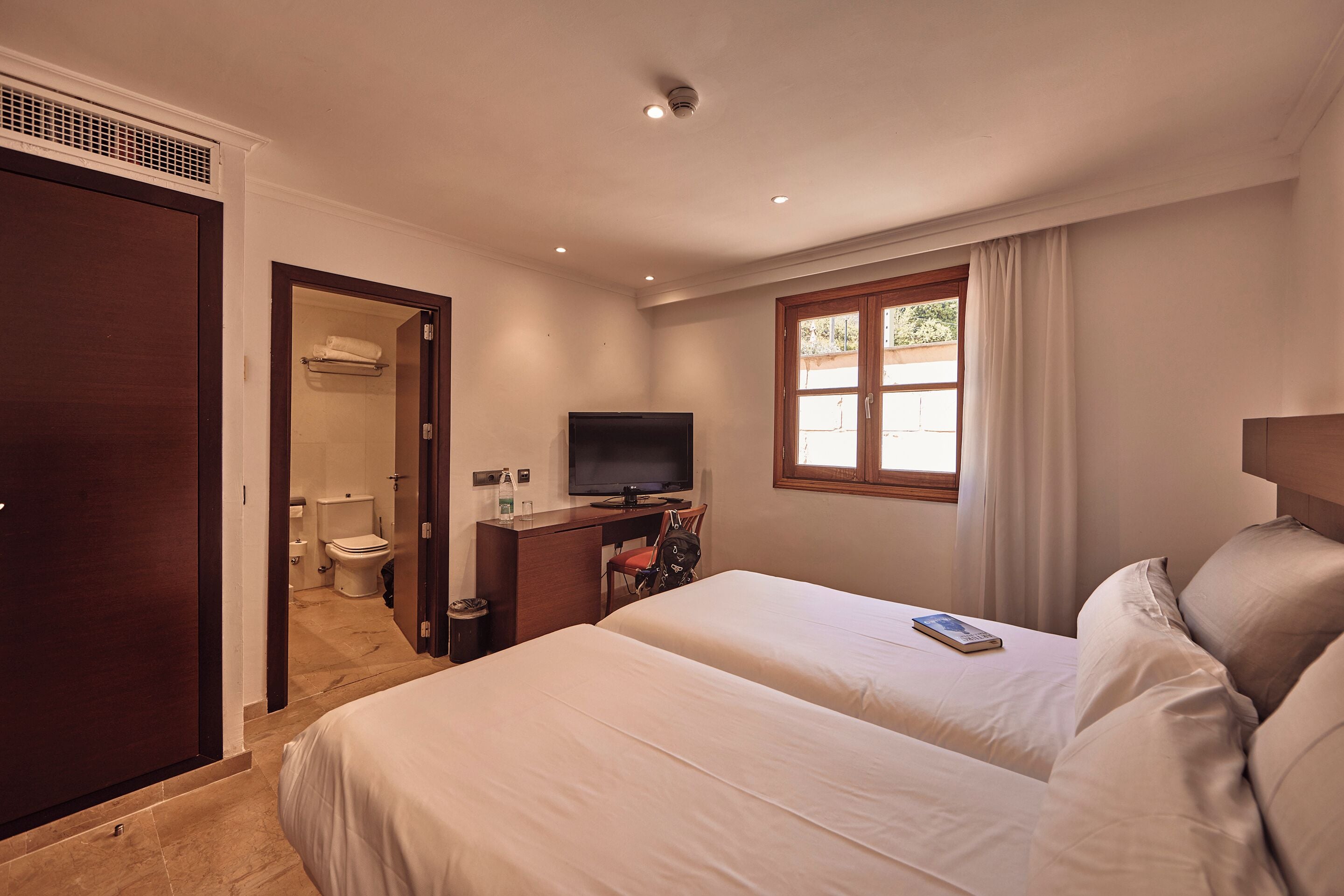 A hotel room with a bed, a desk with a television, a window with curtains, and a bathroom visible through an open door.
