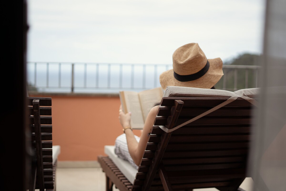 Une personne est assise sur un banc en bois, un livre à la main, et contemple la mer.