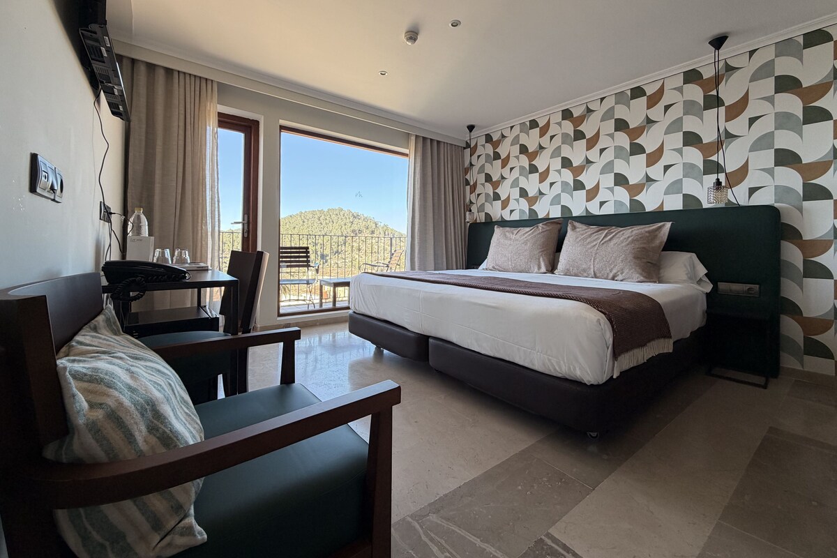 Une chambre d’hôtel équipée d’un lit, d’un bureau, d’une chaise et offrant une vue sur un paysage montagneux grâce à de grandes fenêtres.