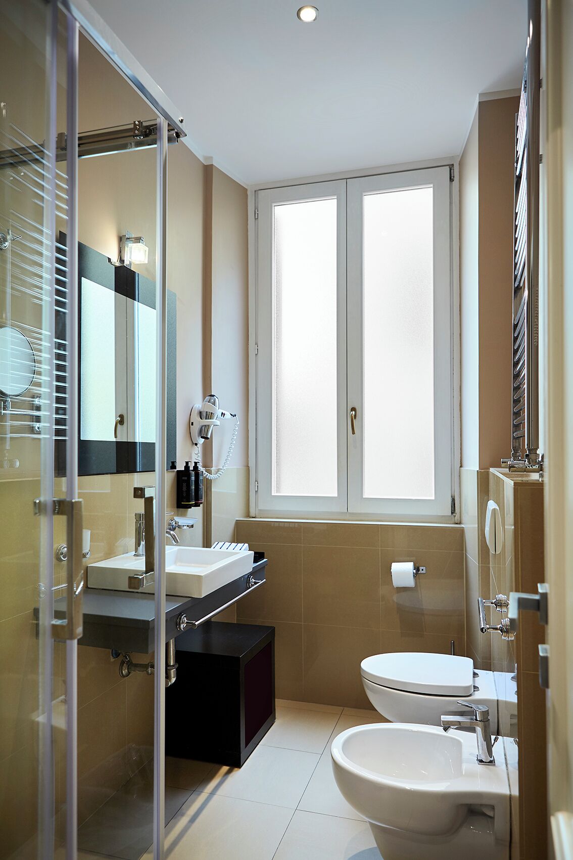 A modern bathroom with a glass shower enclosure, a double vanity with a mirror, a toilet, and a window providing natural light.