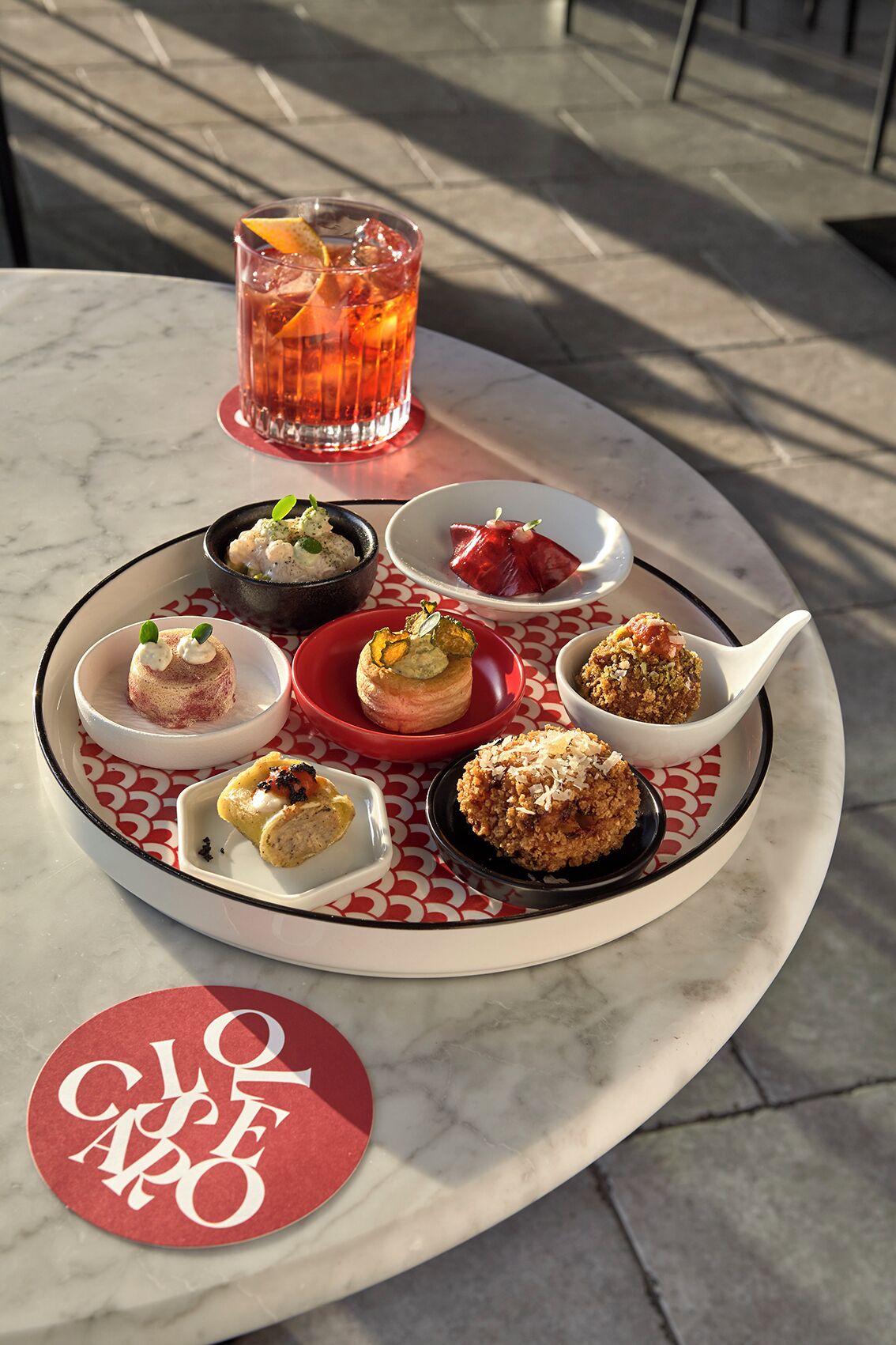 Una mesa con varios platos pequeños y una bebida, al aire libre, con una mesa de mármol y un logotipo en un posavasos.