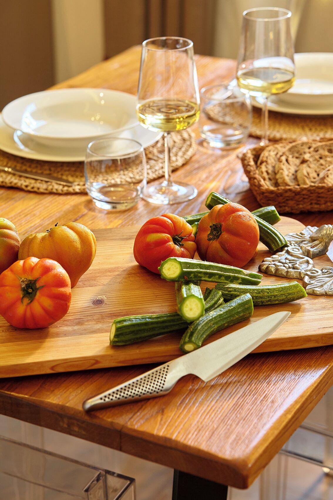 Une table en bois avec une planche à découper, des légumes, un couteau et des verres de vin blanc.