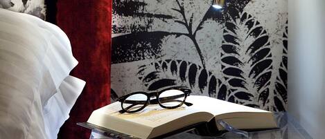 A room with a patterned wallpaper, a red curtain, a clear side table with a book and glasses, and a wall-mounted light fixture.