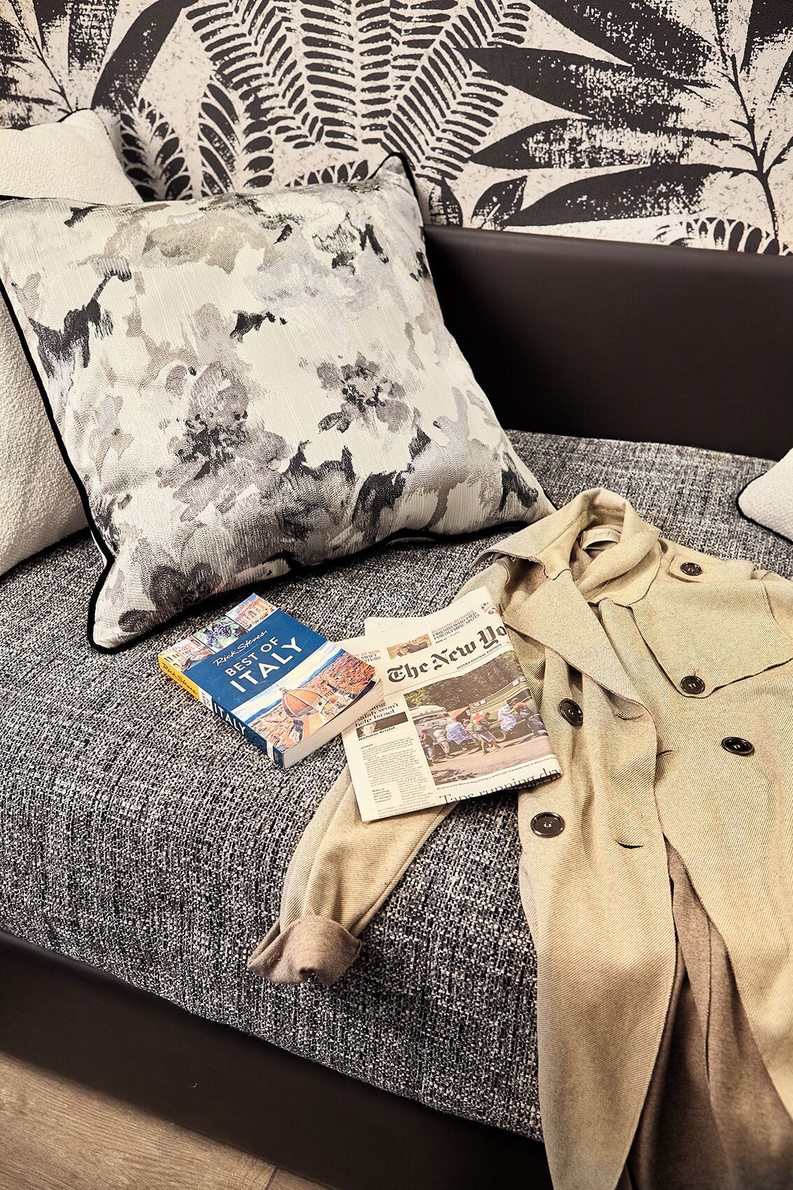 A sofa with patterned cushions, a book titled 'Best of Italy', and a beige coat on a textured fabric.