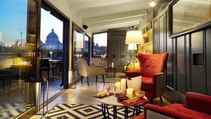 A spacious room with a large window offering a view of a cityscape and a dome building. There are multiple seating areas with a red upholstered chair and a table set with candles and snacks.