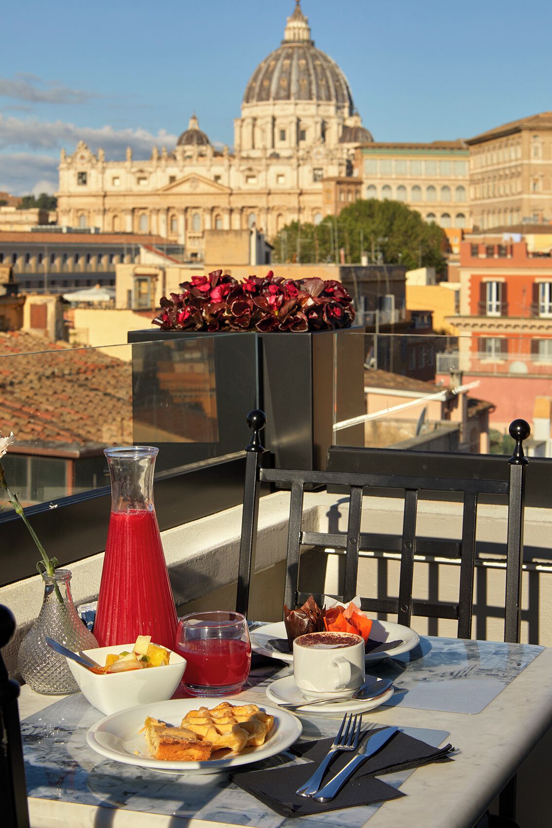 A table with breakfast items, a red drink, and a flower arrangement on a balcony with a view of a large domed building.