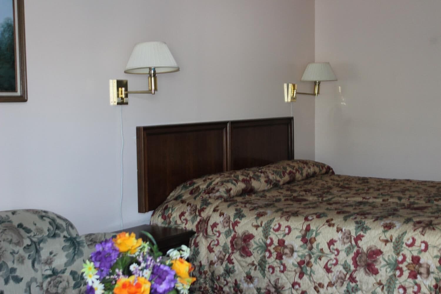 Habitación de hotel con cabecera de madera, dos lámparas de noche, colcha estampada con motivos florales y un arreglo floral sobre la mesita de noche.