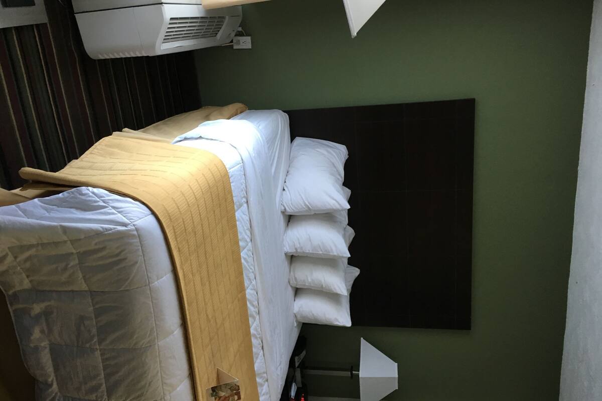 A single bed with a beige blanket and white pillows, a dark headboard, and a wall-mounted air conditioner.