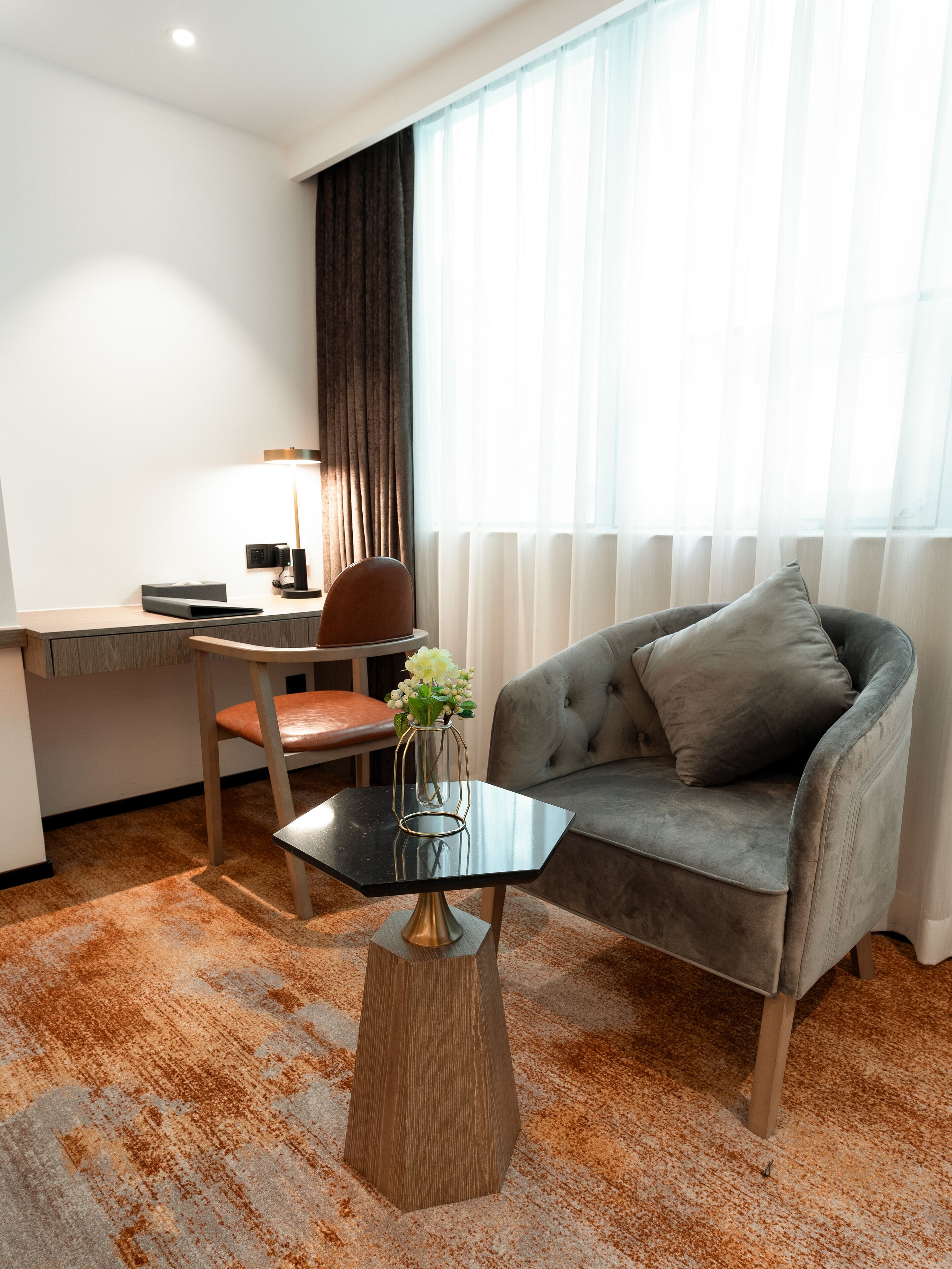 A modern hotel room with a grey sofa, a wooden coffee table with a vase of flowers, a desk with a lamp, and a large window with sheer curtains.