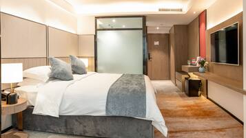 A modern hotel room with a large bed, a bedside table with a lamp, a TV mounted on the wall, and a small seating area with a table and chairs.
