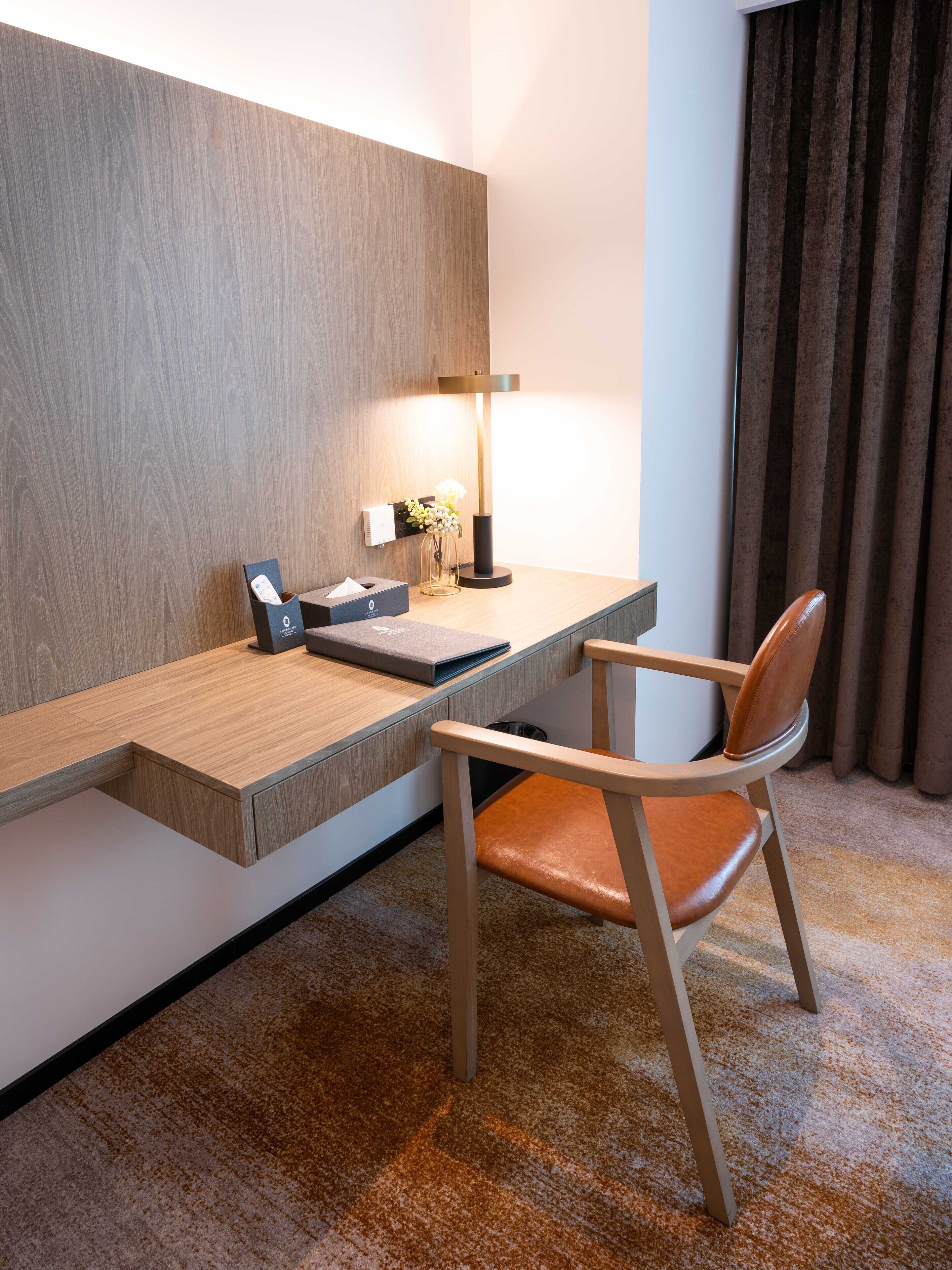 A hotel room with a wooden desk, a chair, a lamp, and a vase with flowers.
