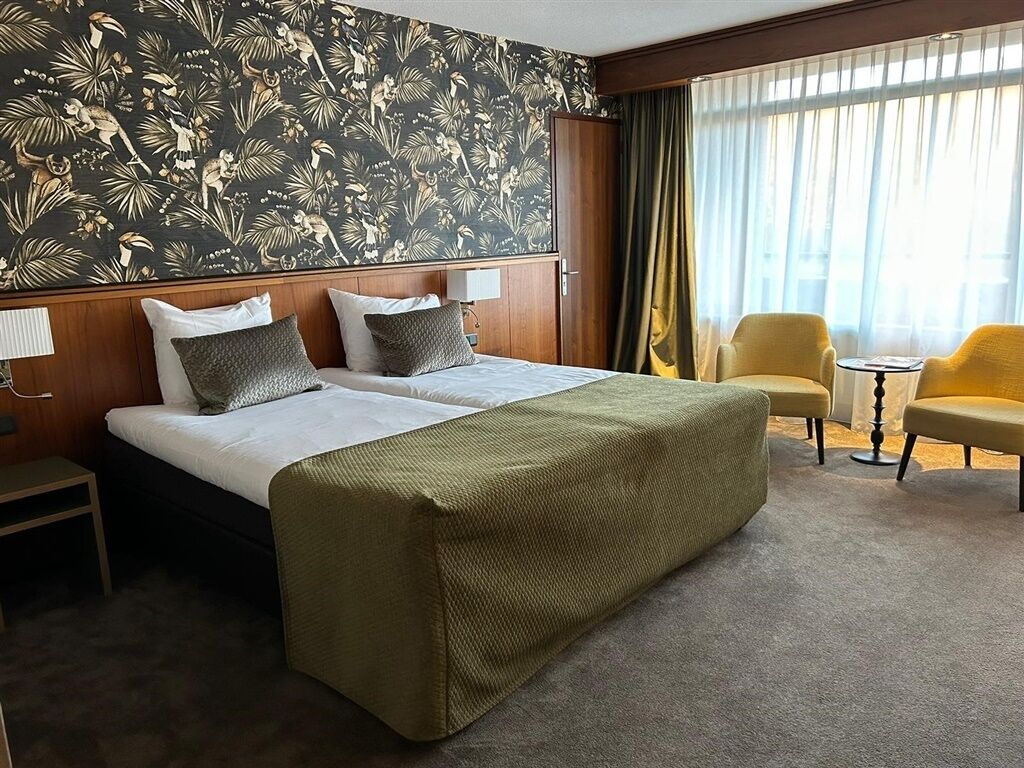 Et hotelværelse med en seng, to sengeborde, en stol, et lille rundt bord og et stort vindue med gardiner.