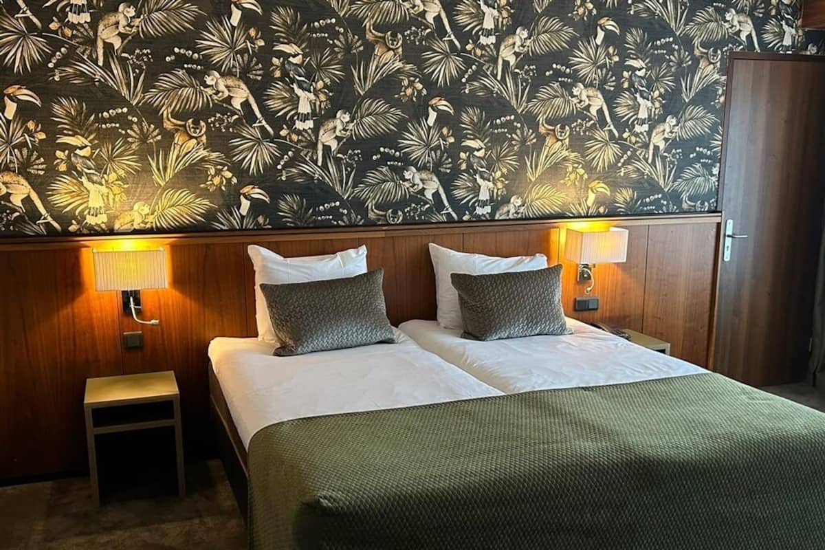 Una habitación de hotel con cama, dos lámparas de noche, una mesita de noche y un papel tapiz floral.