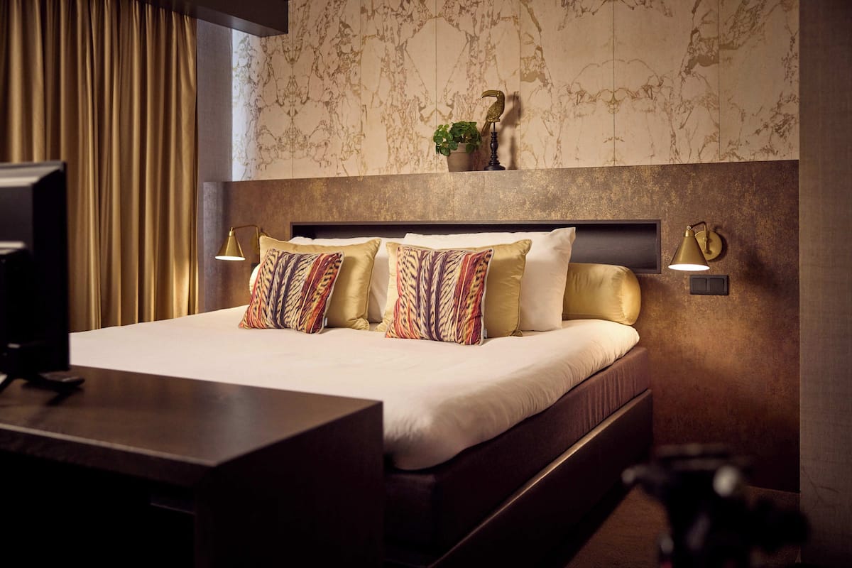 Una habitación de hotel con cama, escritorio, televisor, planta y una pared con diseño tipo mármol.