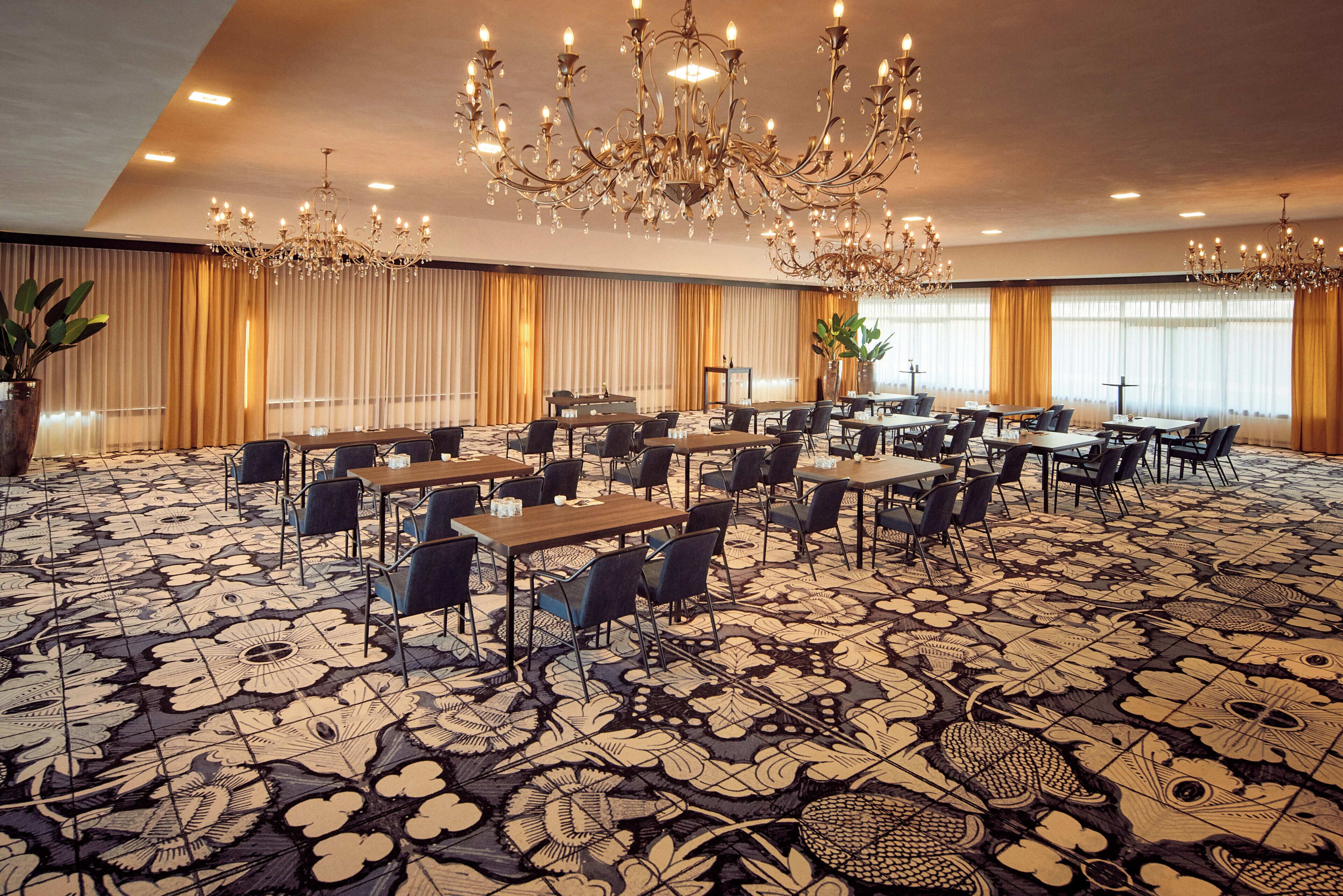 A spacious conference room with a patterned carpet, multiple tables and chairs, and a large chandelier.