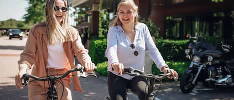 Dos personas andando en bicicleta por un camino pavimentado, con un edificio y motocicletas estacionadas al fondo.