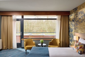 A hotel room with a bed, two armchairs, a small table with a plant, and a view of a brick wall and trees through large windows.