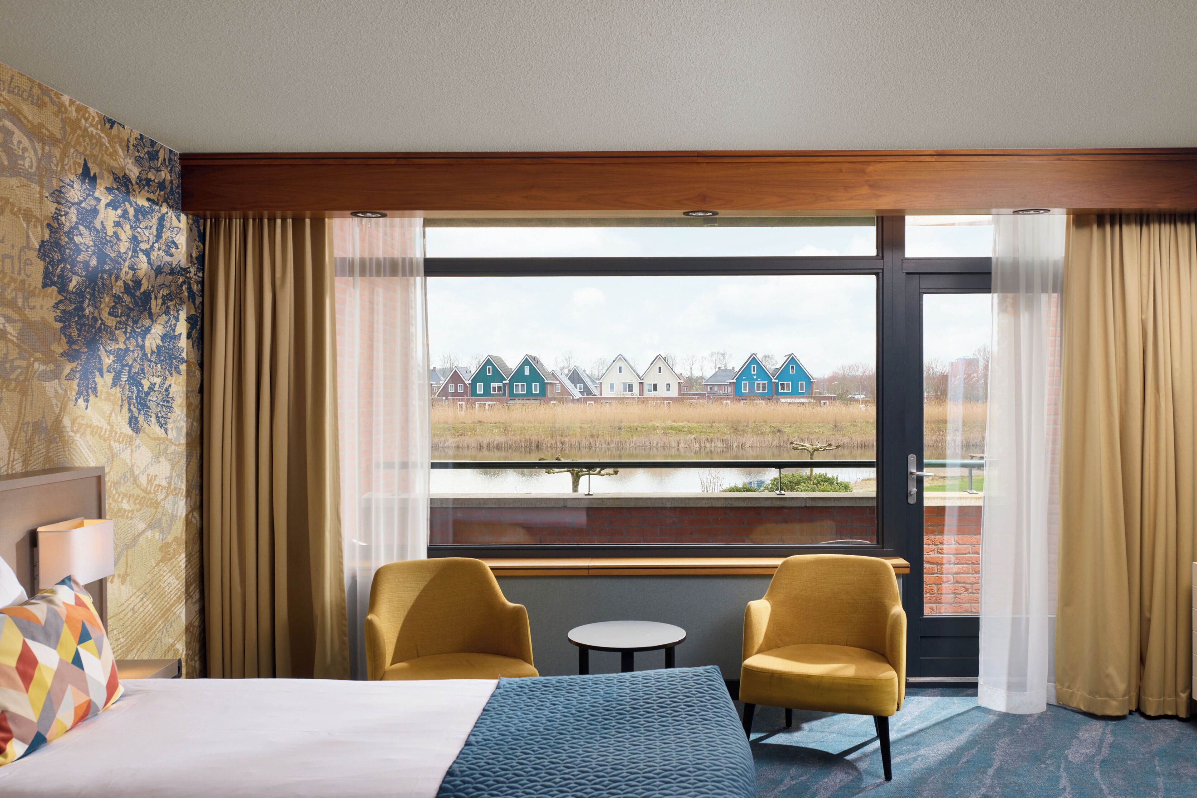 Une chambre d’hôtel avec un lit, deux chaises, une petite table et une grande fenêtre donnant sur des maisons et une étendue d’eau.