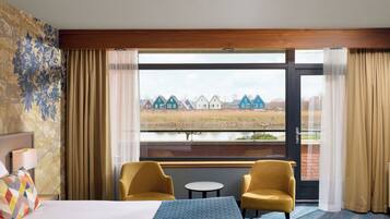 Une chambre d’hôtel avec un lit, deux chaises, une petite table et une grande fenêtre donnant sur des maisons et une étendue d’eau.