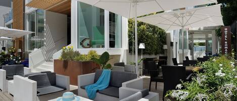 Un área de estar moderna al aire libre con mobiliario de mimbre, un paraguas grande y vistas al interior del edificio.