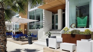 A modern outdoor seating area with white wicker furniture, blue chairs, and a wooden staircase leading to an upper level. - Hotel Romagna (Cattolica)