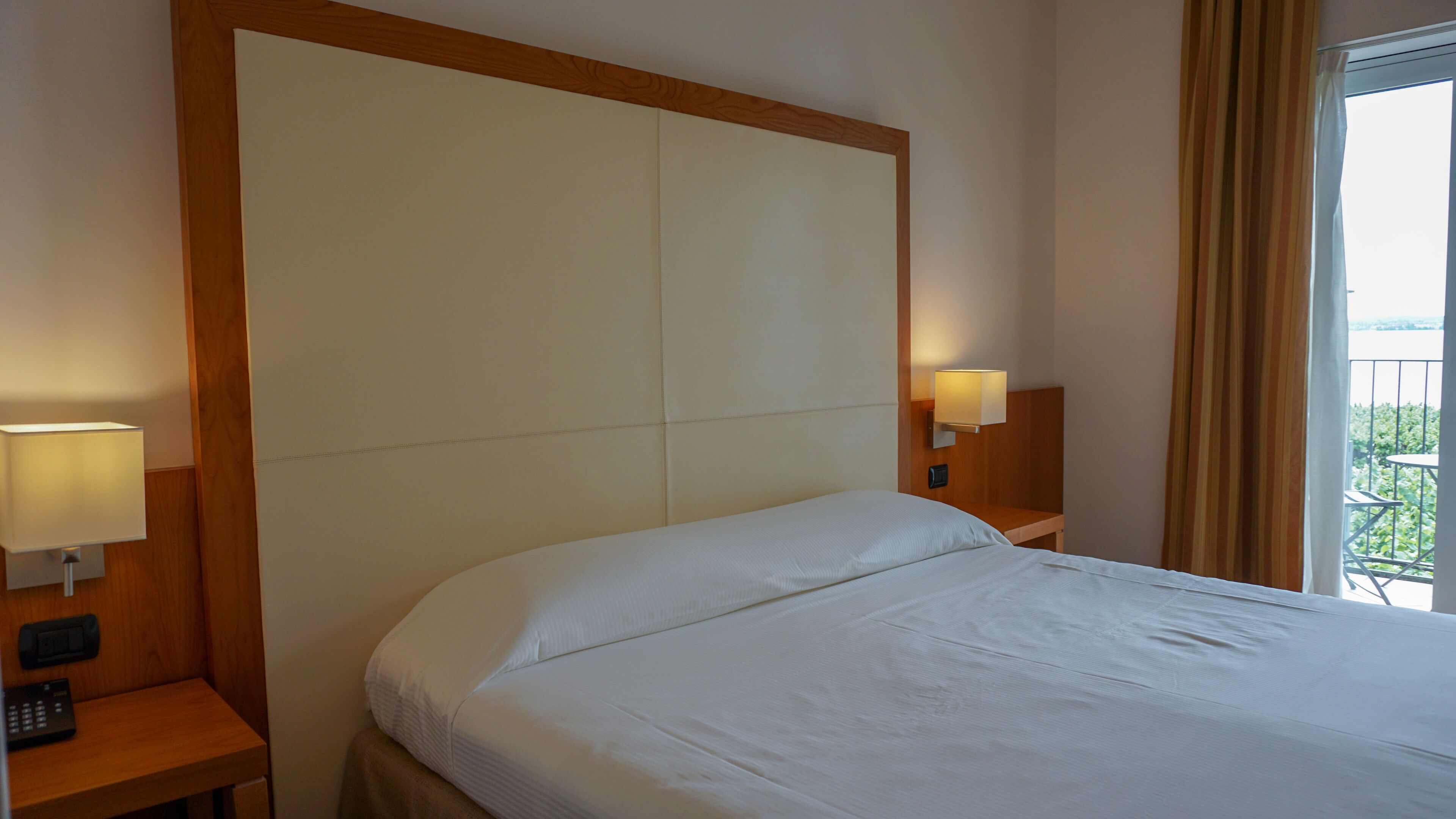 Quarto de hotel com uma cama grande, duas mesas de cabeceira com abajures e vista para o verde pela janela.