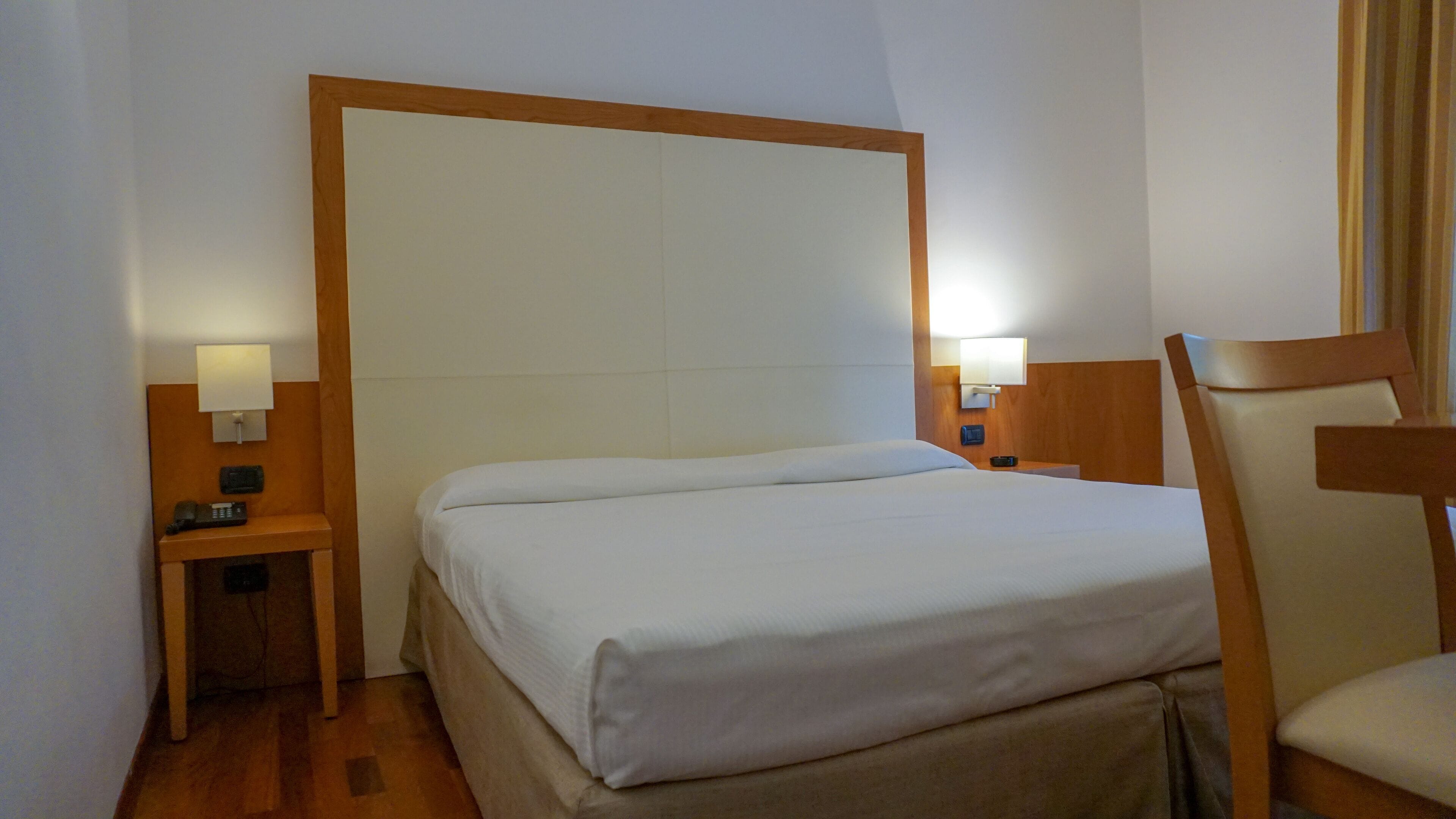 A hotel room with a large bed, wooden headboard, two bedside tables with lamps, a chair, and a small desk with a telephone.