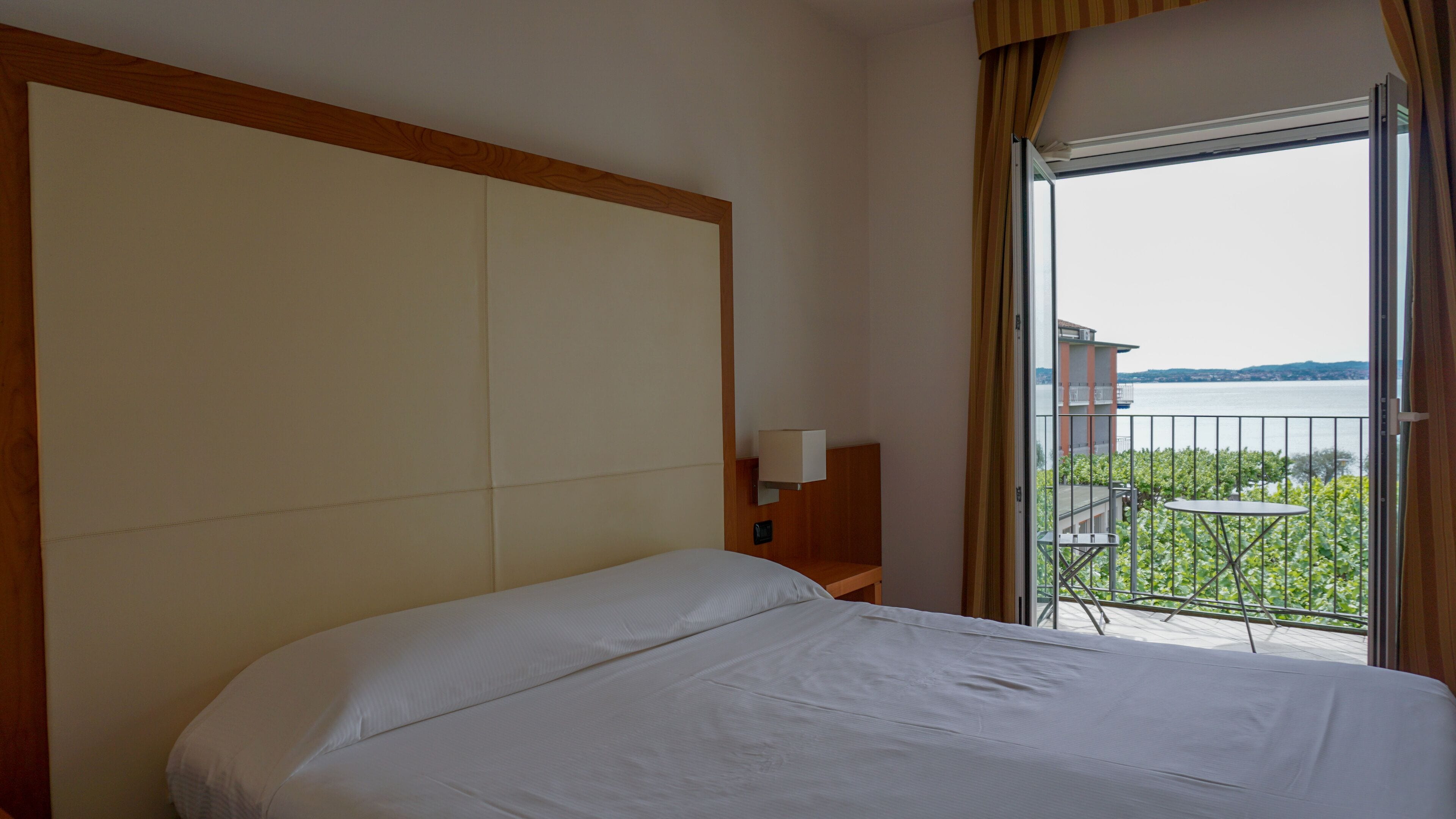 Habitación de hotel con una cama grande, una mesita de noche, una lámpara y vistas a un balcón con mesa y sillas.