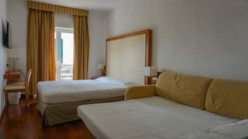 A hotel room with two single beds, a wooden headboard, a sofa, a TV, and a window with curtains.