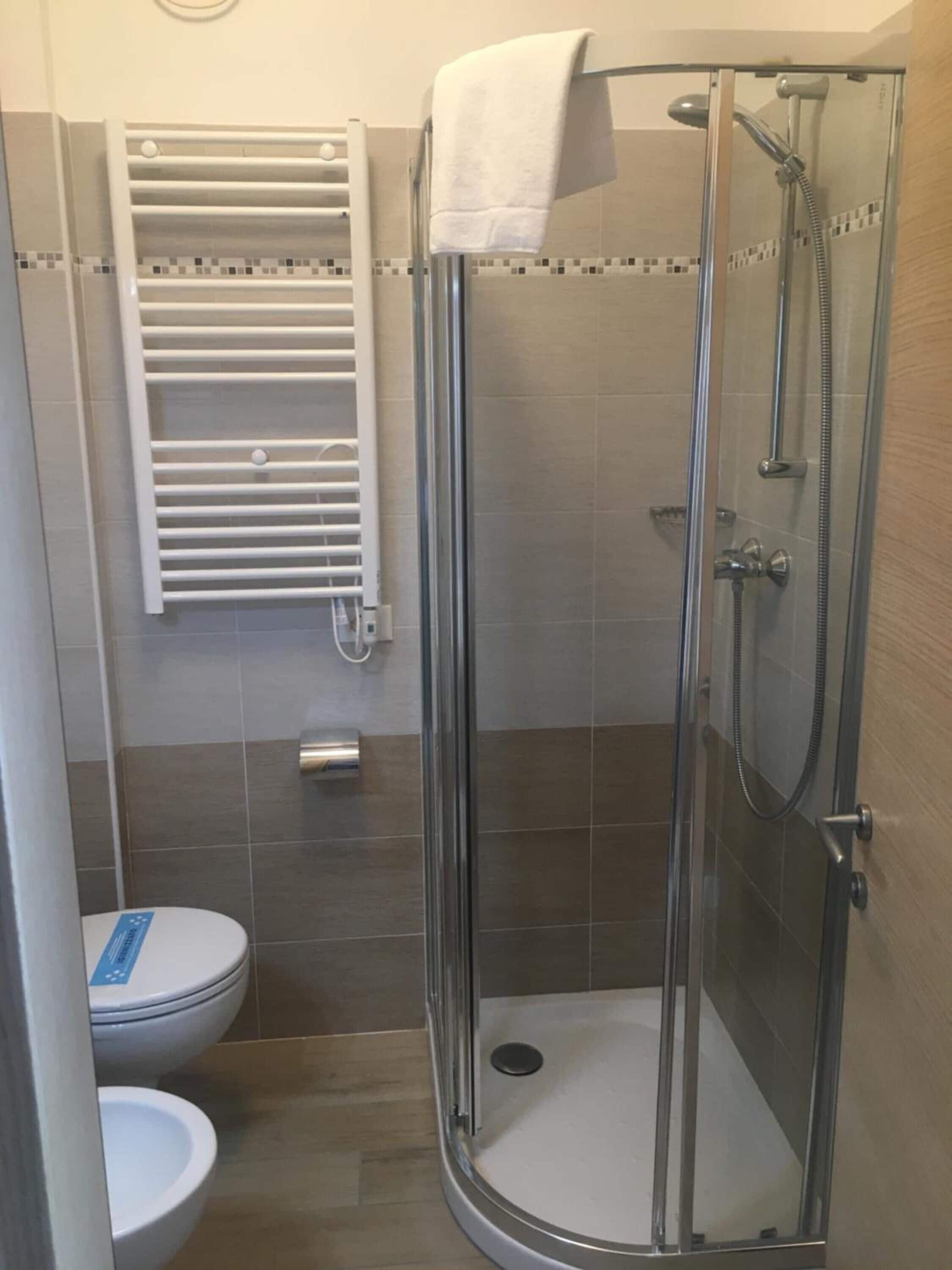 A shower enclosure with a glass door, a white towel hanging on the door handle, and a white toilet with a blue sign.