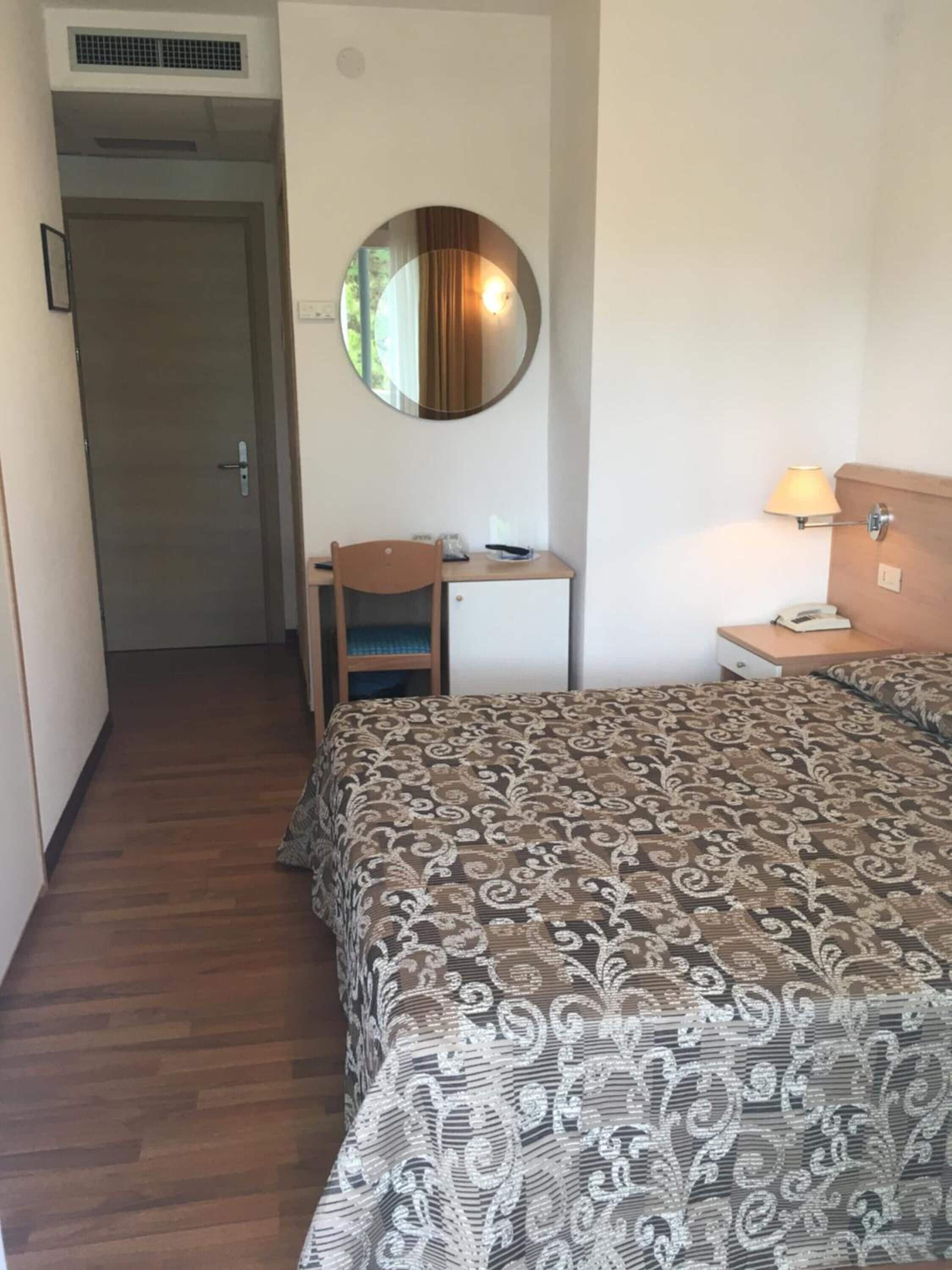 Hotellihuone, jossa on sänky, työpöytä, tuoli, pyöreä peili, lamppu ja puhelin.