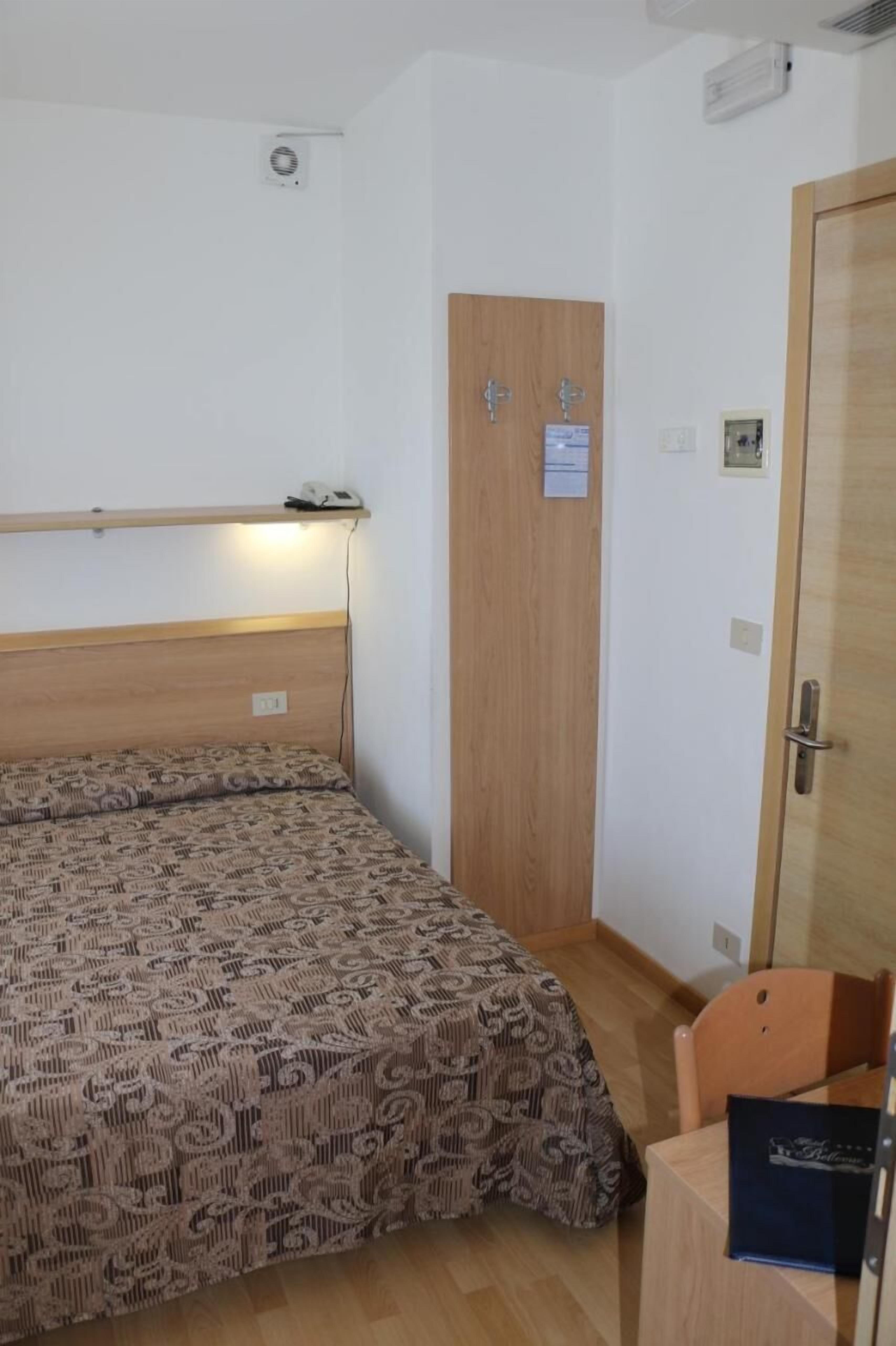 A small, single-room hotel room with a wooden headboard, a bed with patterned bedding, a desk with a chair, and a door with a sign.