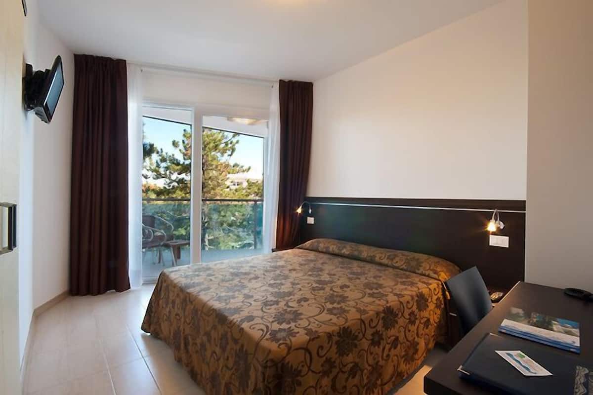Habitación de hotel con cama, escritorio, silla, televisor y balcón con cortinas.