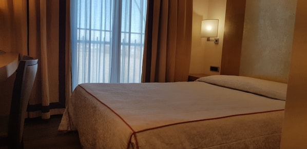 Una habitación de hotel con cama, cortinas, una lámpara de pared y un ventanal con cortinas traslúcidas.