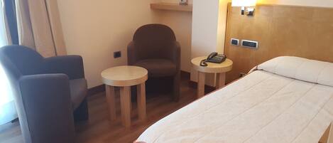 Habitación de hotel con cama, una silla, una mesita con teléfono e iluminación en la pared.