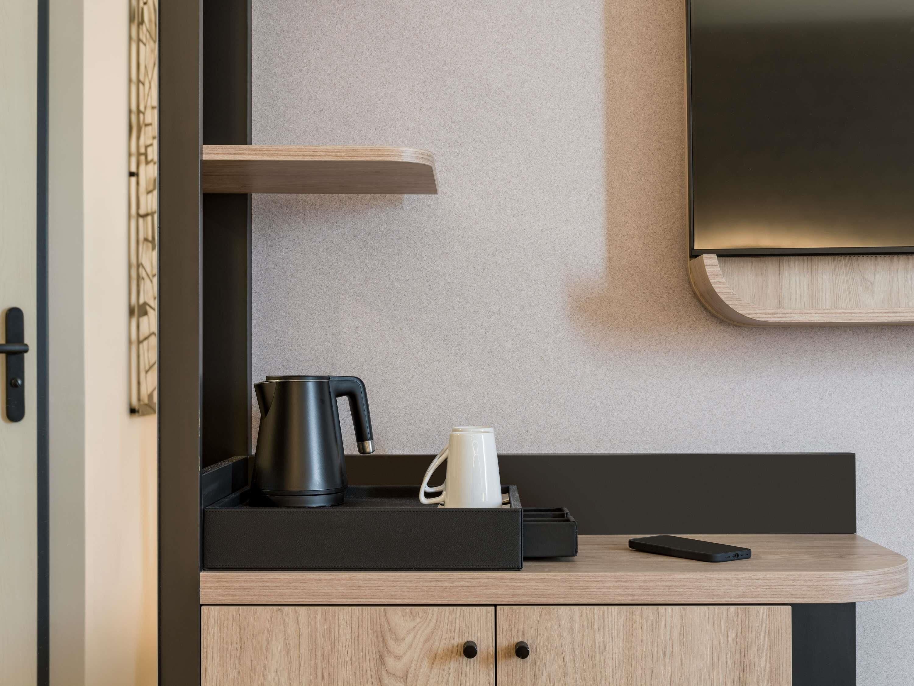 Una kitchenette moderna con una tetera negra, una taza blanca y un teléfono inteligente sobre una encimera de madera.
