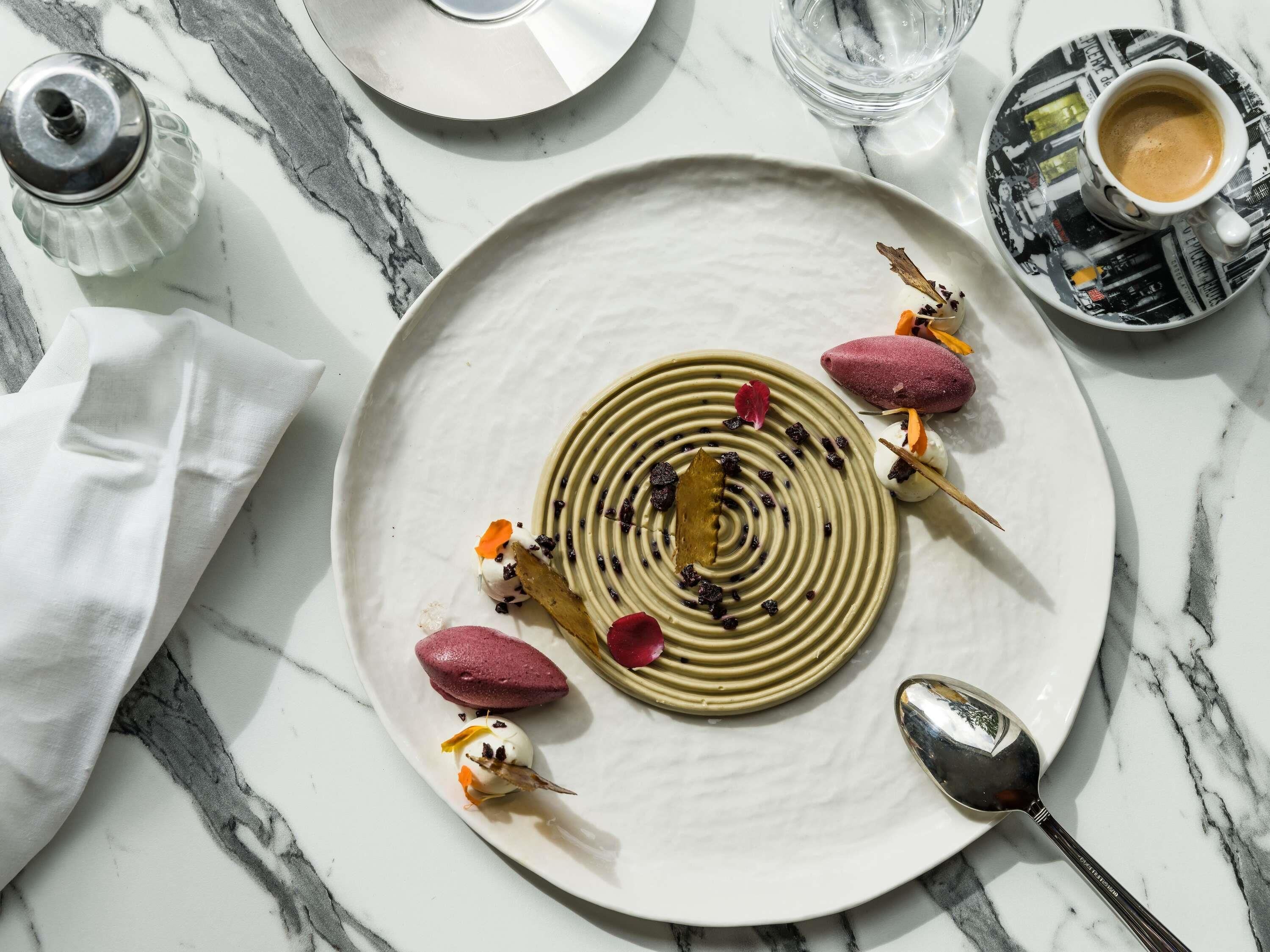 Prato de sobremesa com design espiral, decorado com flores comestíveis e frutas vermelhas, sobre uma mesa de mármore.