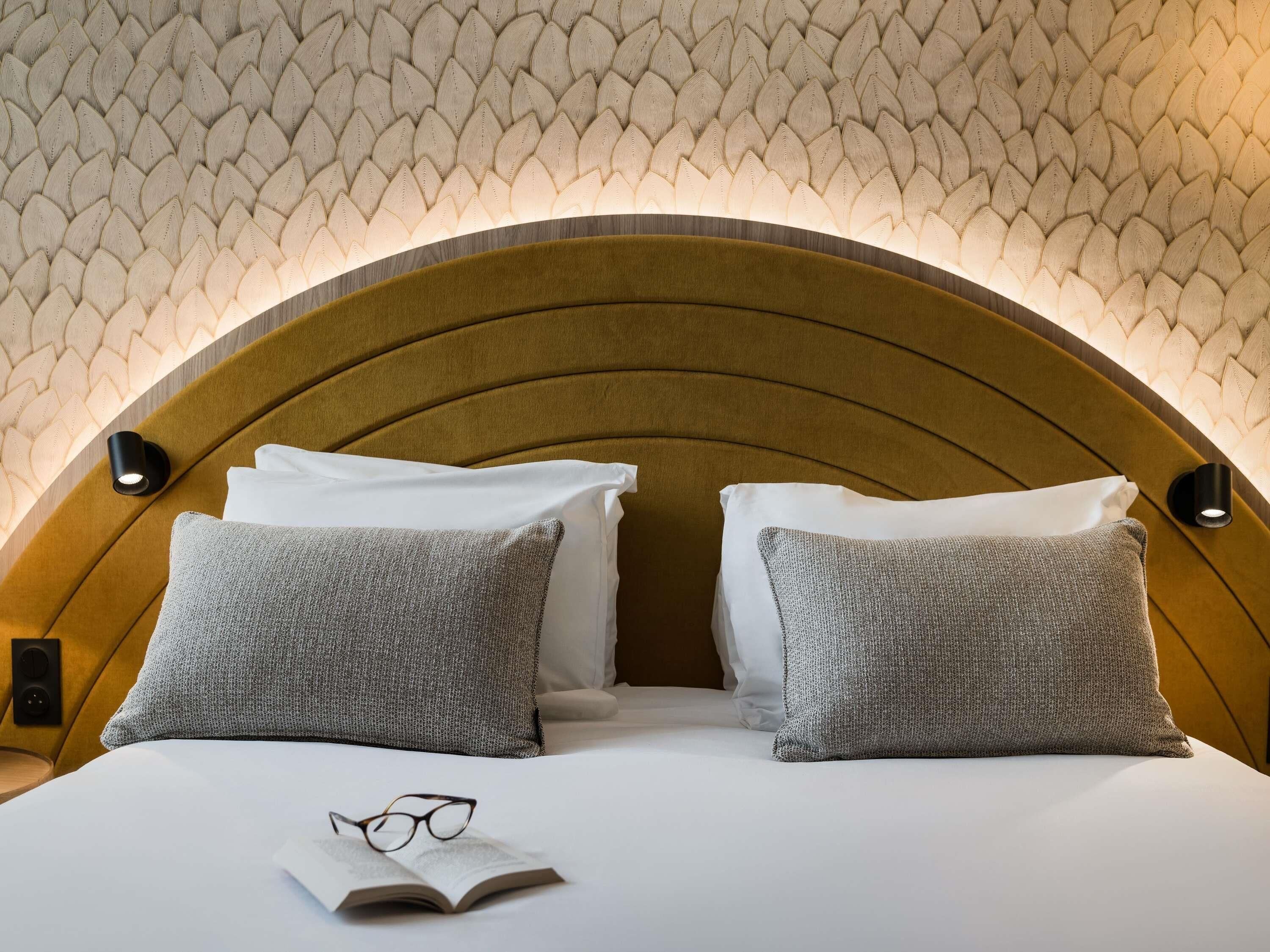 A neatly made bed with white linens, two grey pillows, and a pair of glasses on the bed.