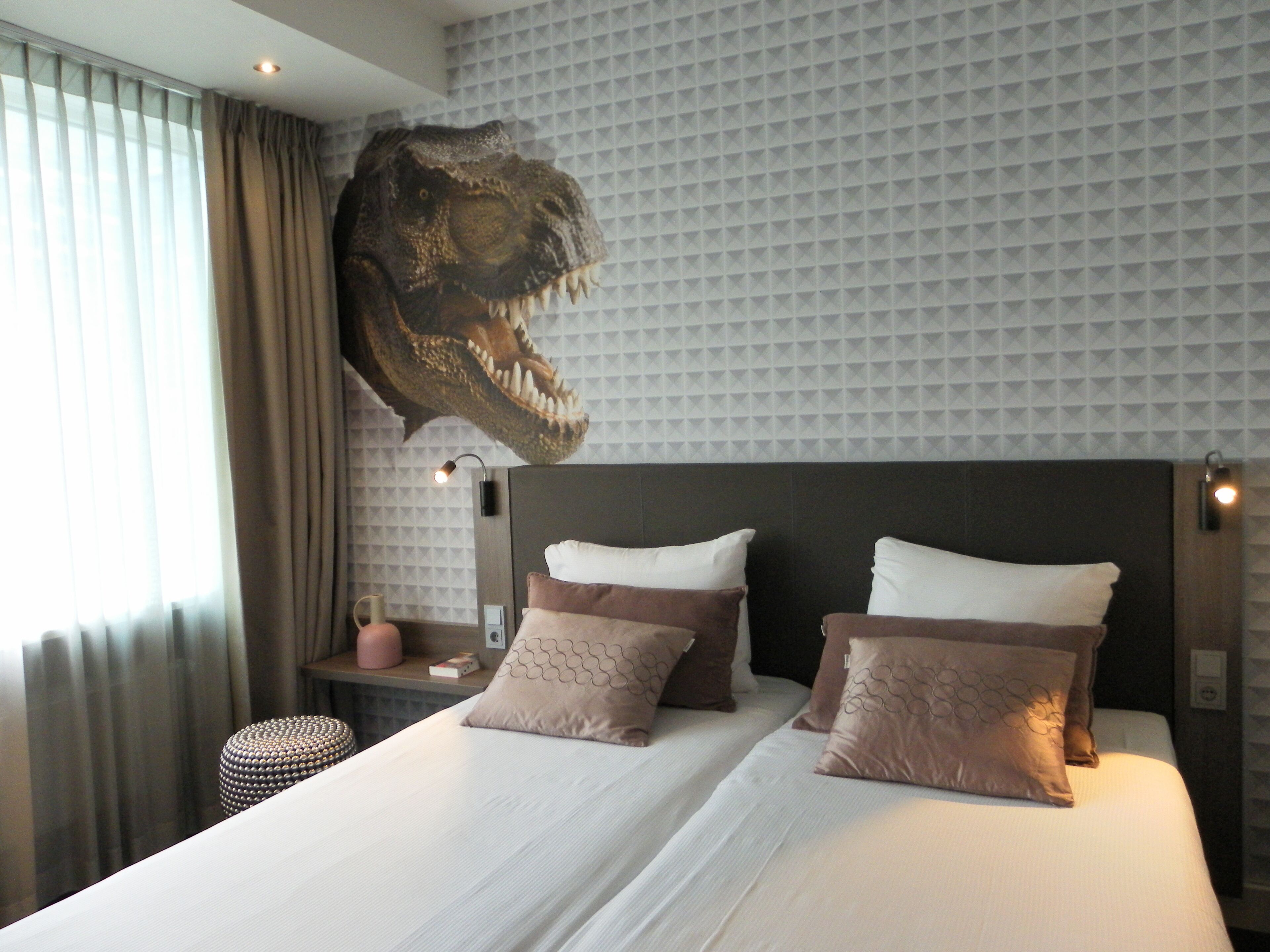 Ein Hotelzimmer mit einer großen Wandmalerei eines Dinosaurierkopfes, einem Bett mit weißer und brauner Bettwäsche sowie einem kleinen Nachttisch mit einer Lampe.