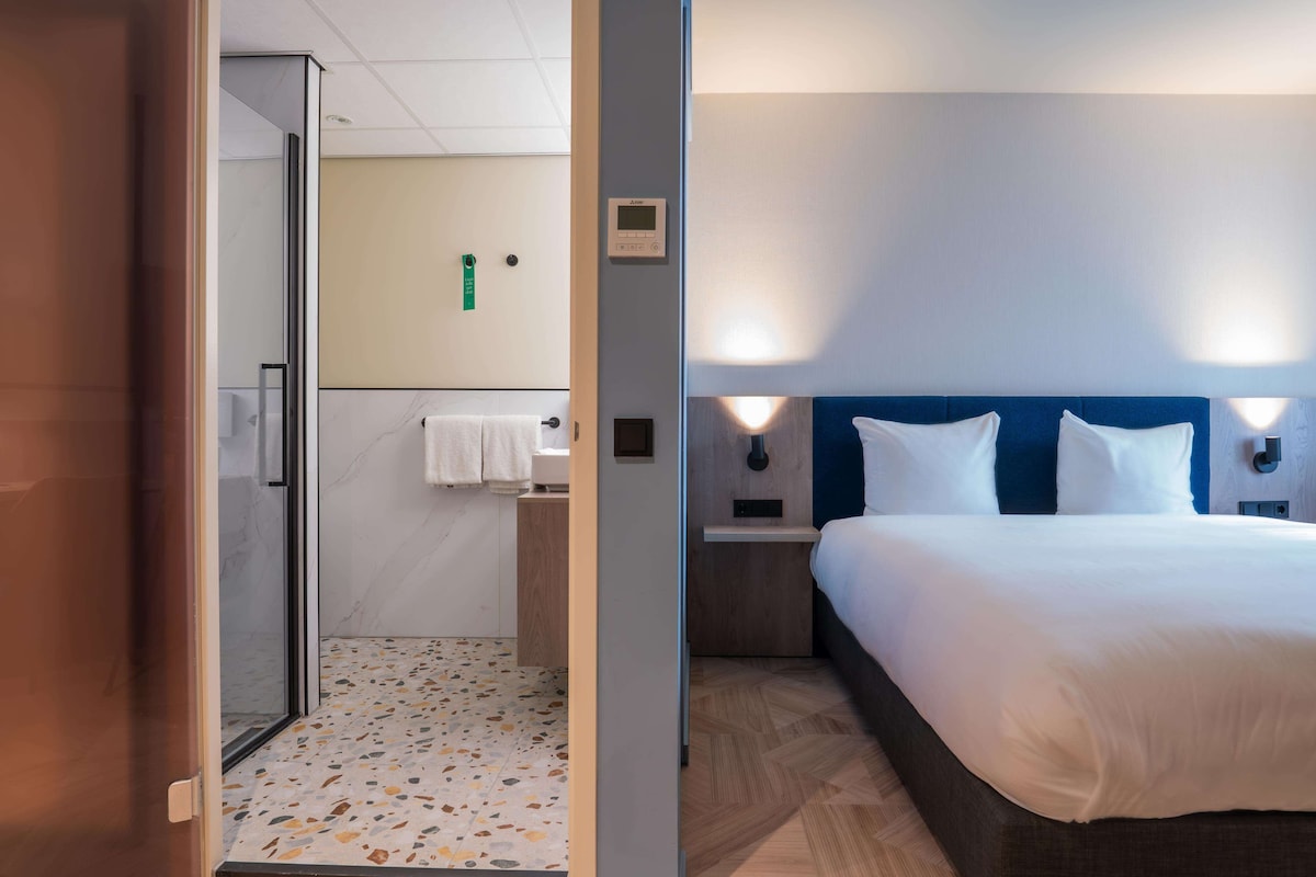 A hotel room with a bed, a nightstand, a wall-mounted light, a shower area, and a bathroom with a towel rack.