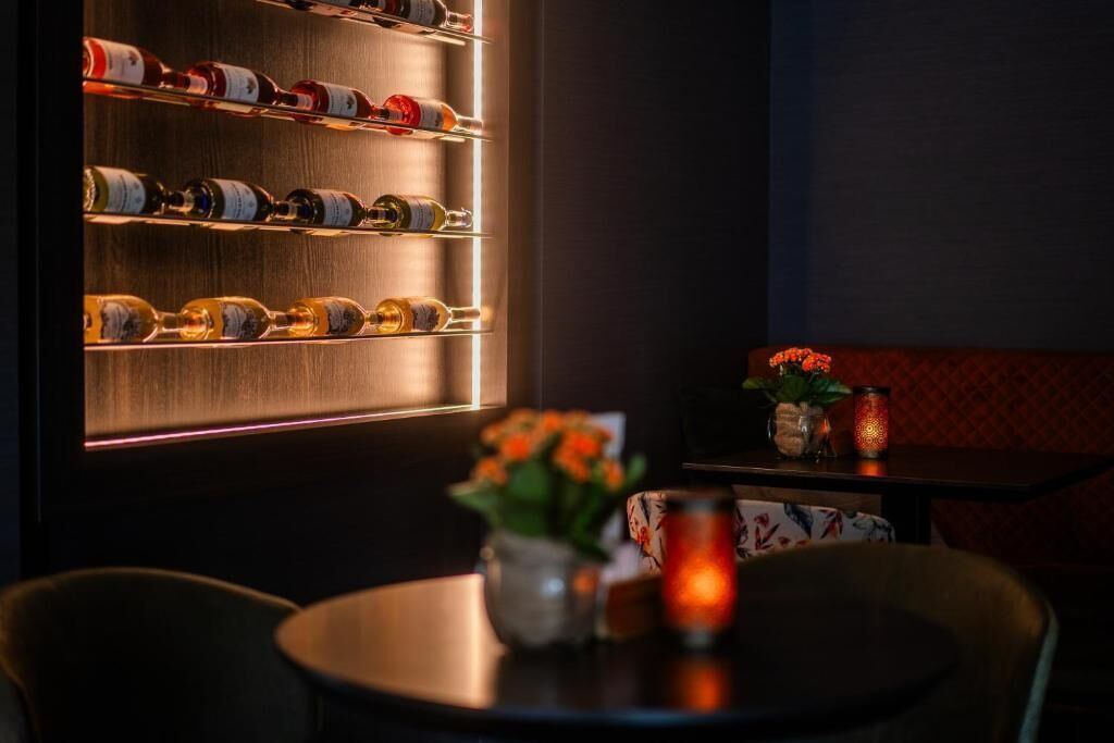 A dimly lit bar area with shelves of bottles, a table with a vase of flowers, and a red cushioned bench.