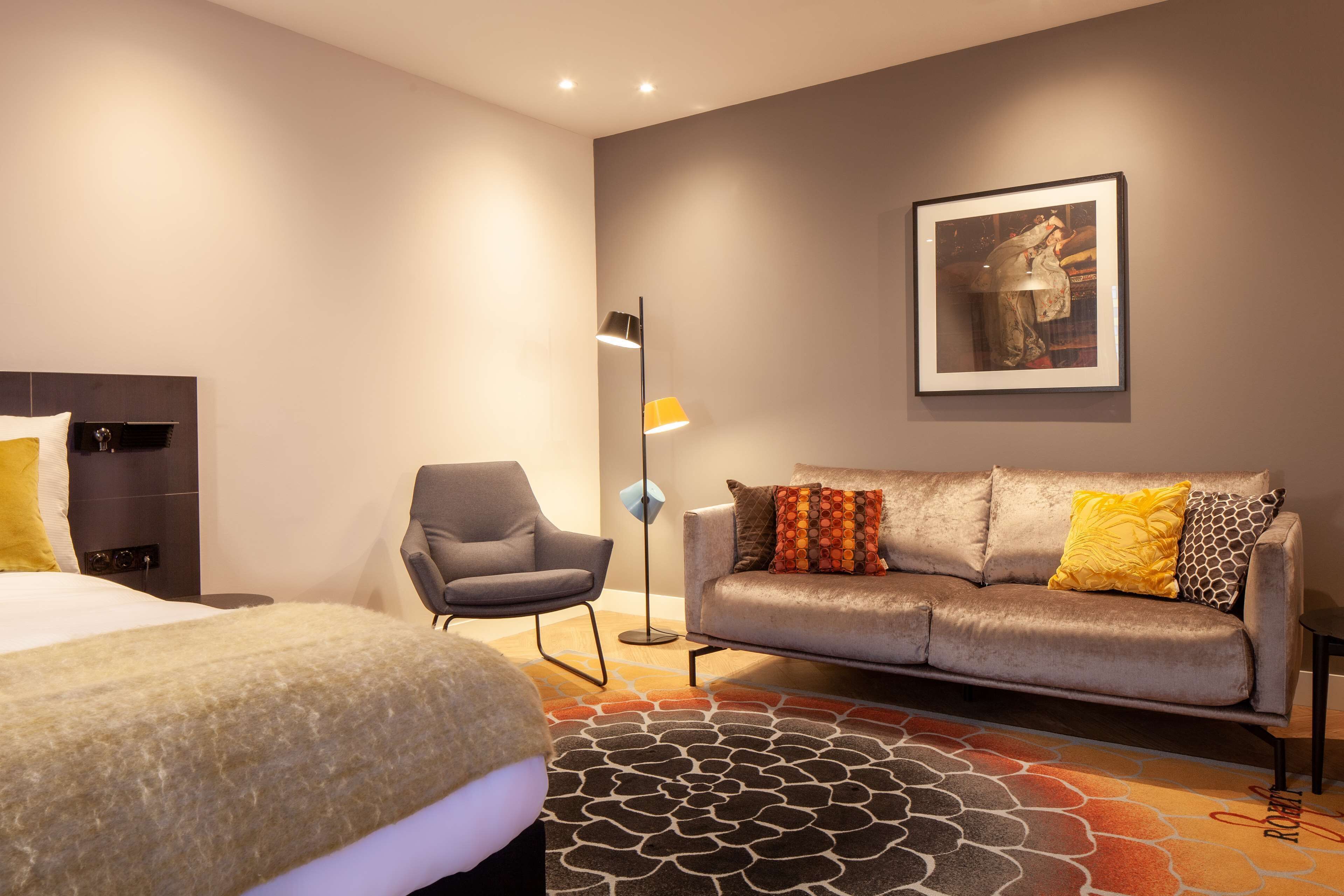 A hotel room with a bed, a sofa, a chair, a floor lamp, and a framed picture on the wall.