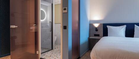Habitación de hotel con cama, mesita de noche, lámpara de pared y puerta que da a un baño.