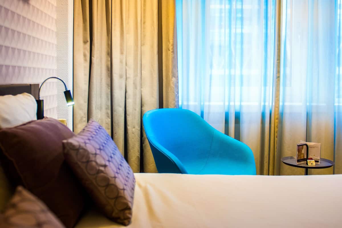 Una camera d'albergo con un letto, una poltrona blu, un tavolino con una lampada e tende.