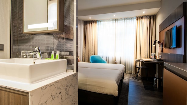 A hotel room with a marble bathroom sink, a bed, a desk, a chair, a TV mounted on the wall, and a window with curtains.