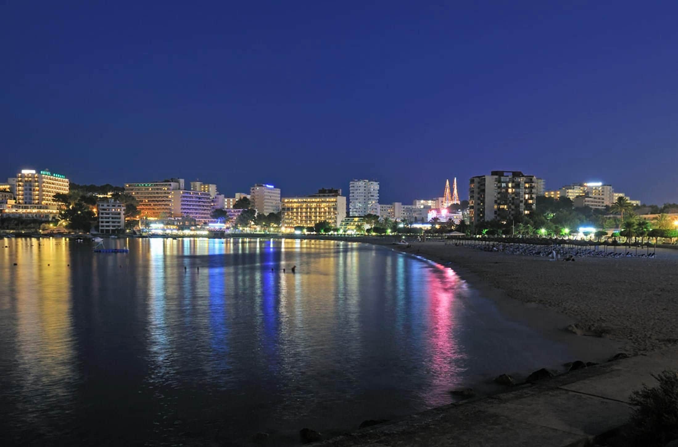Un paisaje urbano costero nocturno con edificios iluminados y un tranquilo paseo marítimo.