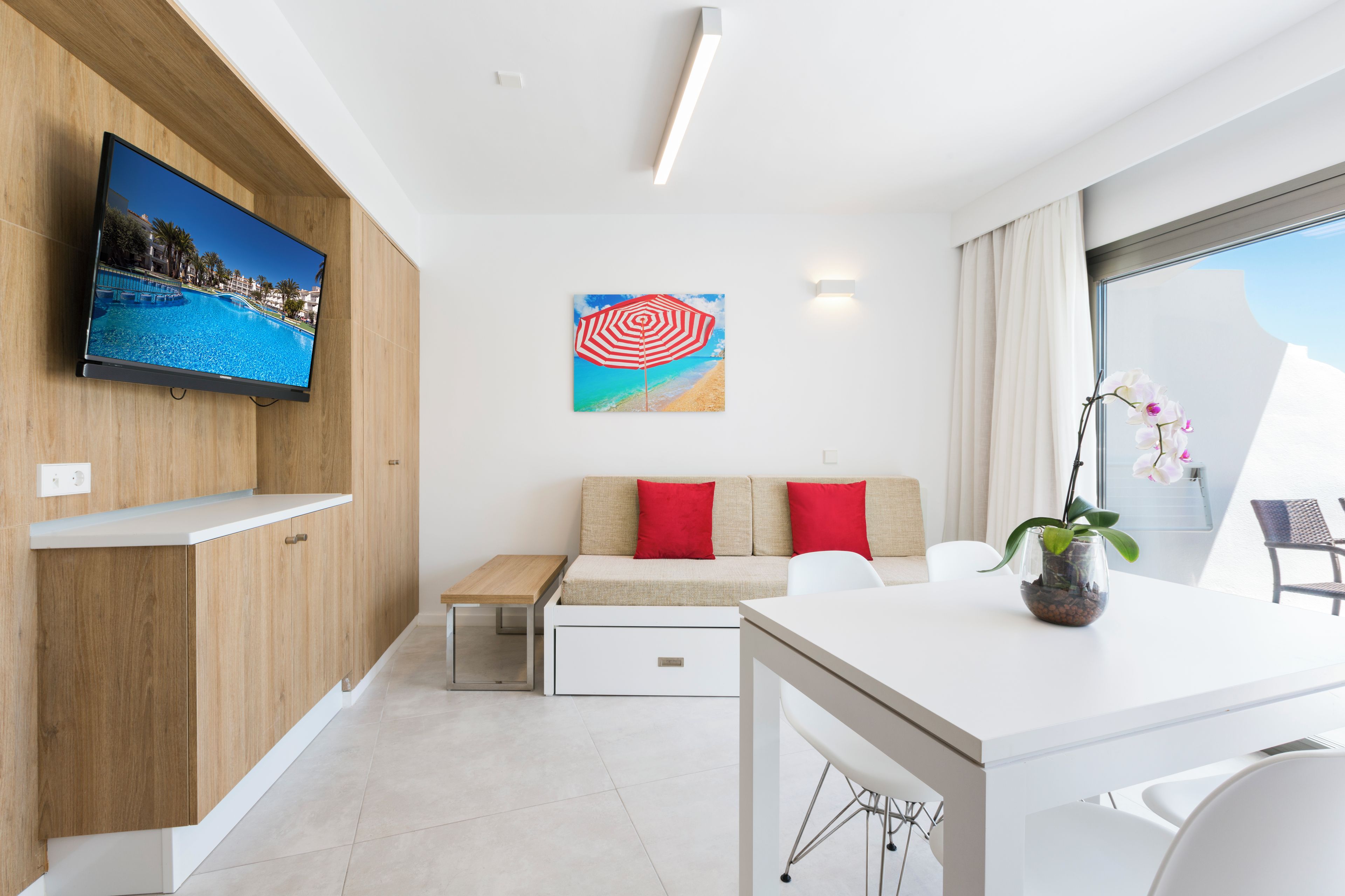 Moderni hotellihuone, jossa on taulutelevisio, rantamaiseman kuvaava taulu, punaisilla koristetyynyillä varustettu sohva, ruokapöytä ja ruukussa oleva orkidea.